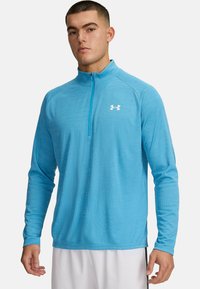 Helles blaues langärmliges Sportshirt mit einem Viertel-Reißverschluss, strukturiertem Stoff und einem kleinen weißen Logo auf der linken Brust.