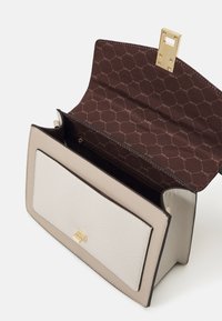 Sac à main structuré beige avec un devant lisse et texturé et une fermeture dorée. L'intérieur présente une doublure marron avec un motif de logo répétitif.