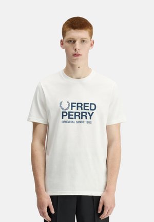 Joven con cabello corto rojo que lleva una camiseta blanca de Fred Perry con texto de logo en azul marino "Fred Perry Original Since 1952" en el frente.
