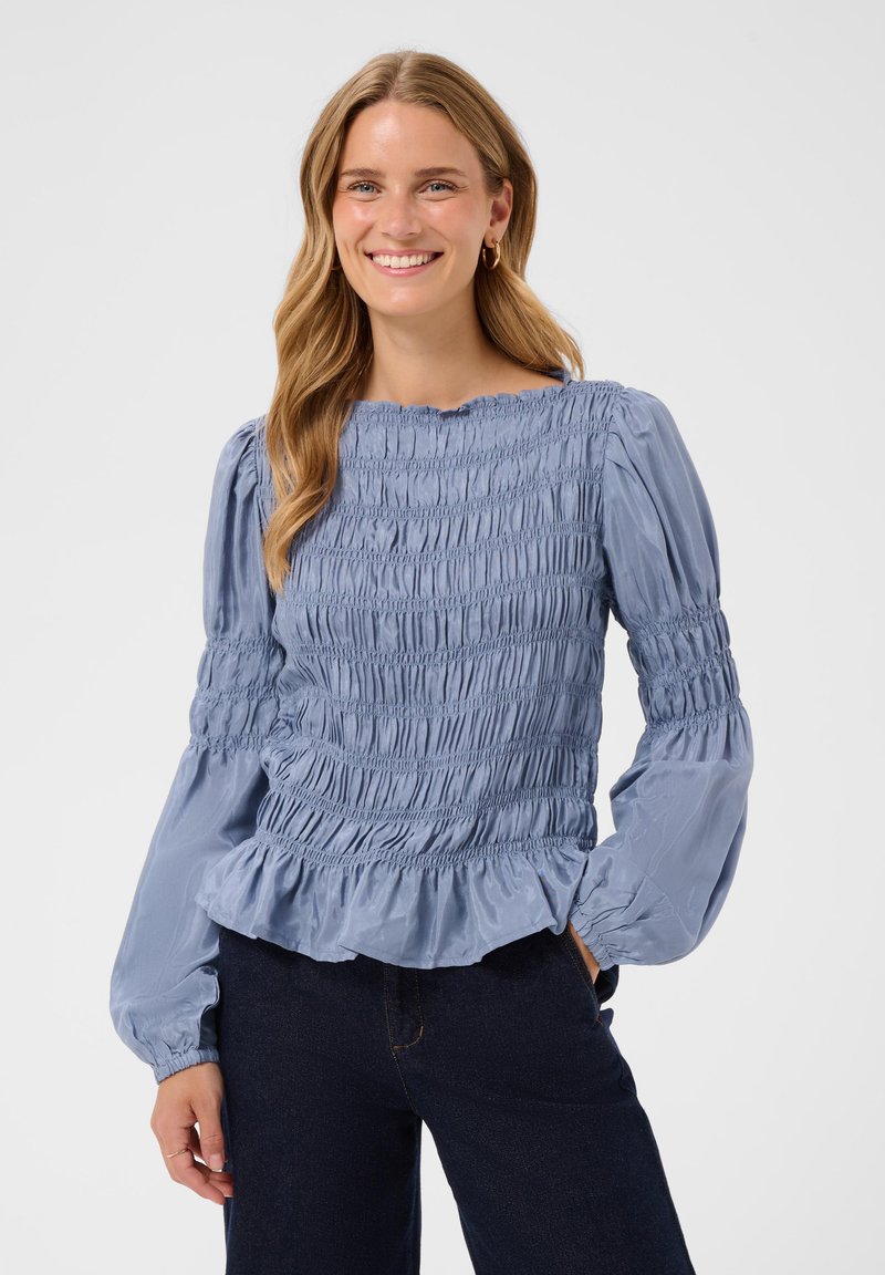 Femme souriante aux longs cheveux blonds portant un chemisier bleu clair froncé avec des manches longues bouffantes et un pantalon foncé sur un fond uni.