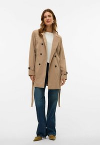 Sieviete stāv, tērpusies bēšā divrindas tranšejas mētelī, baltā krekliņā, zila džinsu biksēs un sinepju dzeltenās norādījuma kurpēs uz vienkārša baltā fona.