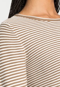 Camicia a righe beige e bianca, con scollatura tonda e tessuto strutturato. Primo piano della spalla mostra sottili righe alternate.