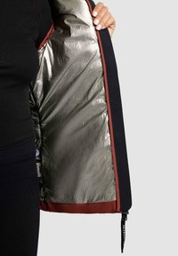 Black jacket with a shiny silver lining and a maroon trim. Features a zipper closure and a tag at the bottom. Textured fabric visible.