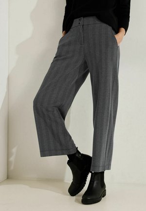 Kobieta ubrana w krótkie szare spodnie w jodełkę, czarne botki Chelsea i czarną bluzkę, stojąca z rękami w kieszeniach na tle jednolitej ściany.