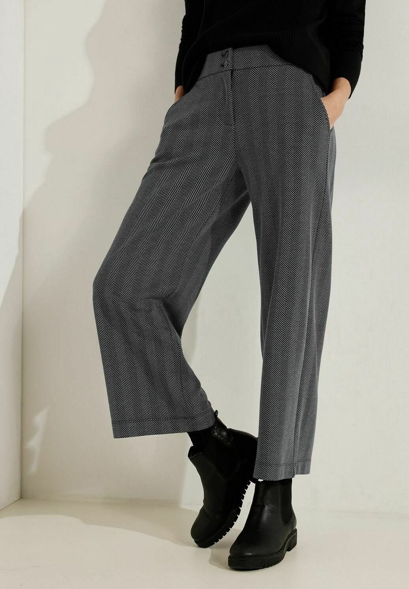 Femme portant un pantalon gris à chevrons raccourci, des bottines Chelsea noires et un haut noir, les mains dans les poches, devant un mur uni.