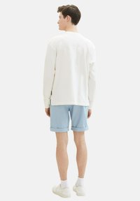 Camisa blanca de manga larga con un patrón texturizado, combinada con pantalones cortos de color azul claro enrollados en los bordes. El modelo lleva zapatillas deportivas blancas.