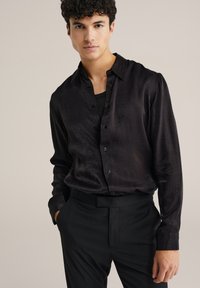 Mees, kellel on seljas läikiv must nööbid särk musta alussärgi üle, mustade pükstega, seismas ühtlase heleda tausta ees.