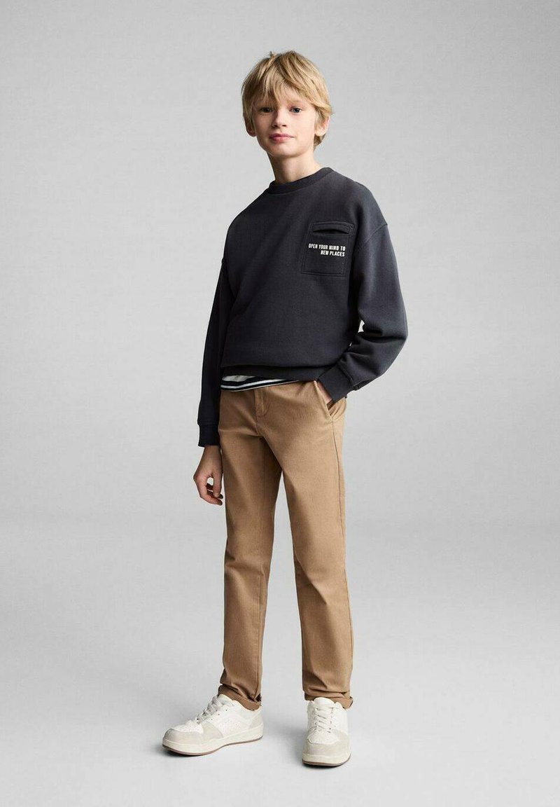 Siva majica s kapuco in sprednjim žepom, rjavi hlače in bele superge. Majica ima napis na žepu in udoben kroj.