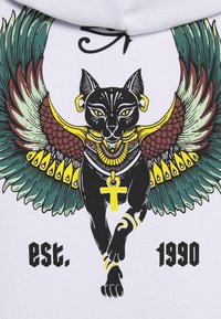 Gatto nero con occhi gialli e gioielli in stile egiziano, grandi ali colorate, che tiene un ankh, con il testo "fondato nel 1990" su sfondo bianco.