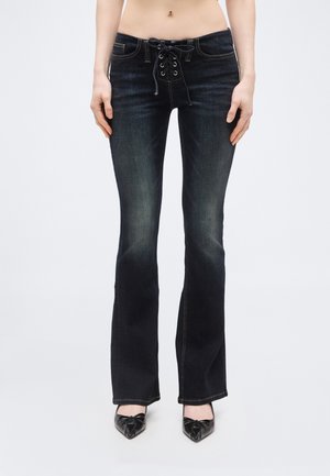 Γυναίκα που φοράει μαύρο τζιν με κορδόνια και καμπάνα στο κάτω μέρος και μαύρες γόβες με μυτερή μύτη, στέκεται μπροστά σε απλό φόντο.