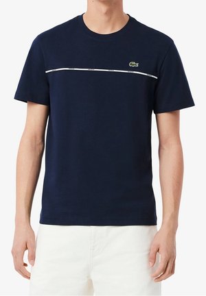 Uomo che indossa una maglietta blu navy a maniche corte con una striscia orizzontale bianca e un piccolo logo verde di coccodrillo sul petto.