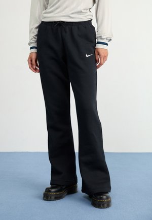 Persoon die zwarte Nike-sweatpants, een wit shirt met lange mouwen en gestreepte manchetten, en zwarte platformlaarzen draagt, staand op een blauwe vloer.
