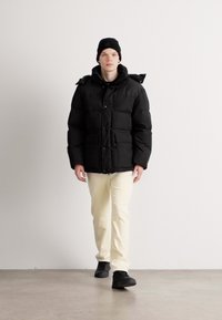 Black puffer jacket with a shiny texture, large front pockets, and a hood. Paired with cream-colored pants and black shoes.