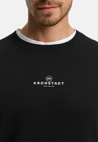 Sudadera negra con textura acanalada, con un logo blanco "KRONSTADT" y la frase "CASUAL SINCE 1999" impresos en el frente. Cuello blanco en capas.