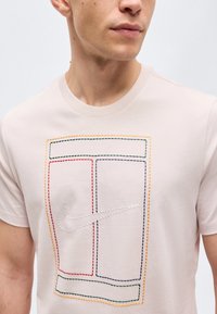 Homme portant un t-shirt beige clair avec des formes rectangulaires multicolores cousues et un logo Nike swoosh blanc cousu sur la poitrine.