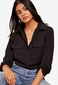 Camisa negra de botones con dos bolsillos en el pecho, mangas remangadas y cuello tipo solapa, combinada con jeans de mezclilla azul de tiro alto.