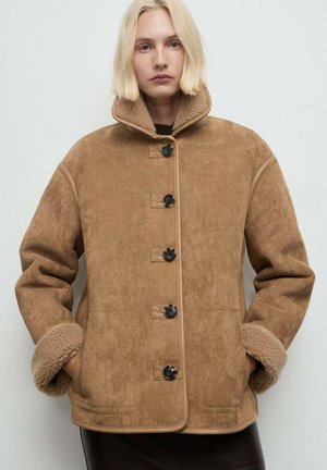 Chaqueta de ante color beige con un cuello alto, que presenta cinco botones negros, puños de borreguillo y una superficie texturizada. Diseño casual y oversize.