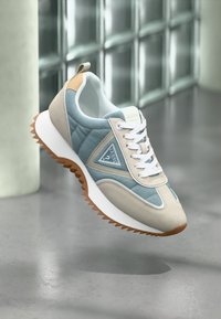 Beige og lyseblå Guess-sneaker med hvide snørebånd og brun rillede sål, svævende over en skinnende grå gulv og væg af glasblokke.