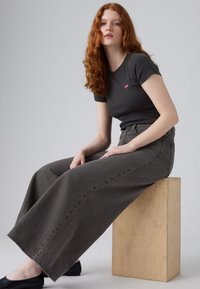 Grijze wijde spijkerbroek van denim, gecombineerd met een getailleerd zwart T-shirt met een klein rood logo, gedragen met zwarte ballerina's.