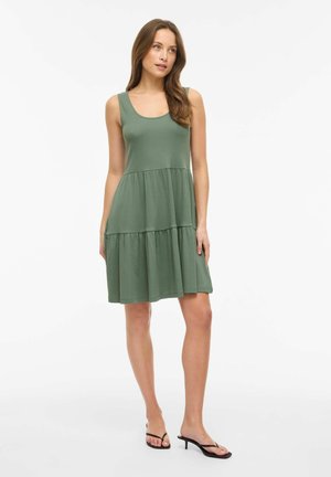 Femme portant une robe olive à étages sans manches et des sandales noires à talons, debout devant un fond blanc uni.