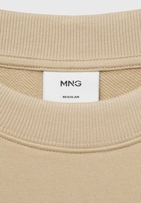 Beige bavlněná mikina s žebrovaným kulatým výstřihem. Štítek ukazuje značku "MNG" a velikost "Regular." Textura je hladká s jemným povrchem.