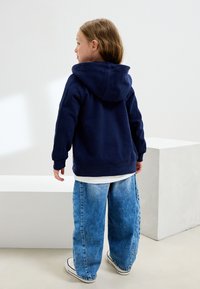 Sweat à capuche bleu marine avec une finition texturée, associé à un jean en denim bleu clair oversize et des baskets blanches, vu de dos.
