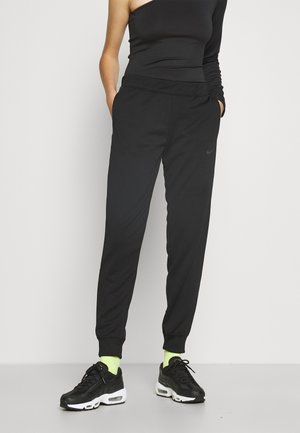 Person trägt schwarze Jogginghosen, ein schwarzes Top mit einem Ärmel, neongelbe Socken und schwarze Turnschuhe und steht mit den Händen in den Taschen.