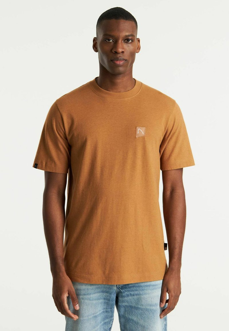 Braun Baumwoll-T-Shirt mit Rundhalsausschnitt, kurzen Ärmeln und einem kleinen Logo auf der linken Brust. Kombiniert mit hellen Jeans.