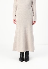Beige gulvlengde skjørt laget av mykt strikket stoff med ribbet midjebånd, kombinert med svarte ankelstøvler. Har en rett, flytende silhuett.