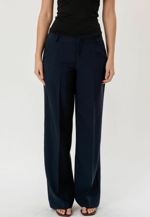 Nő sötétkék, széles szárú nadrágban, fekete testhezálló felsőben és barna, nyitott orrú szandálban, egy egyszerű világos háttér előtt állva.