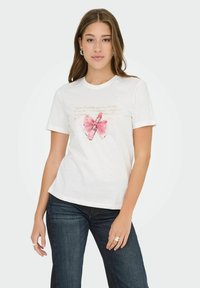 Jonge vrouw in een wit T-shirt met een grafiek van roze lippenstift en een strik, gecombineerd met donkerblauwe jeans, staande tegen een lichte achtergrond.