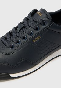 Dunkelblaues Leder-Sneaker mit strukturierten Akzenten, weißer Kontrastsohle und goldfarbener "BOSS"-Markierung an der Seite mit perforierten Details.