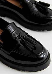 Mocasines de charol negro con punta redonda, borlas decorativas y suela de goma gruesa con detalles texturizados.