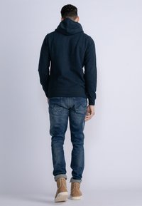Marineblauwe hoodie met een ontspannen pasvorm, gestructureerde stof en een voorzak, gecombineerd met lichtgewassen denimjeans en tan suède laarzen.