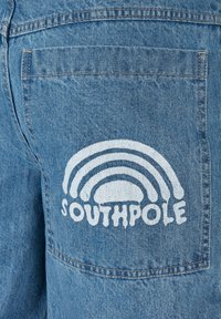 Denim-housut, joissa on takatasku, jossa on valkoinen graafinen kuvasto sateenkaaresta ja sana "SOUTHPole" sen alapuolella, sinisen farkkukankaan taustalla.