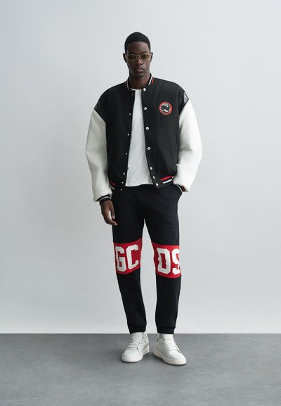 Varsity jacket in black with white sleeves, paired with black pants featuring red accents and white letters. White sneakers complete the look.