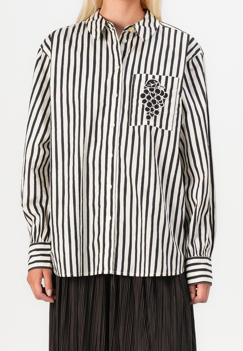 Femme portant une chemise à rayures verticales noires et blanches avec boutons et broderie de grappe de raisin sur la poche poitrine gauche.