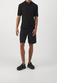 Černý pletený polo svetr s krátkým rukávem, nošený s černými džíny šortkami a černými slip-on botami. Zřetelné textury a vzory viditelné.