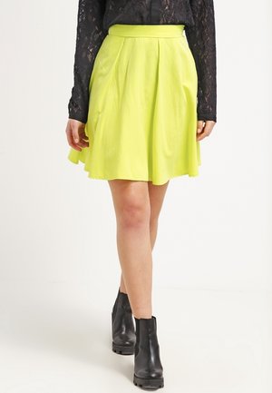 Mujer con falda plisada de color amarillo brillante, top de encaje negro de manga larga y botines negros, de pie sobre un fondo blanco liso.