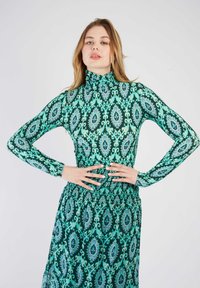 Robe verte à manches longues avec des motifs floraux bleus et violets complexes, col haut et détail de taille froncé. Tissu lisse et léger.