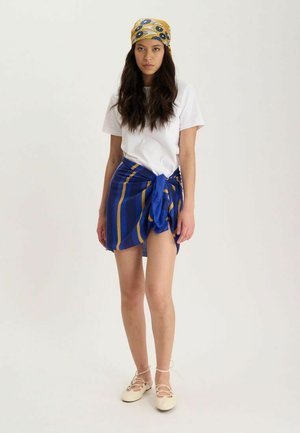 Jeune femme aux longs cheveux foncés portant un t-shirt blanc, une jupe rayée bleue et jaune nouée à la taille, un foulard fleuri et des chaussures à lacets couleur crème.