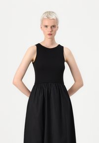 Μαύρο φόρεμα με εφαρμοστό αμάνικο τοπ και φαρδιά φούστα. Το ύφασμα έχει λεία υφή, με λεπτομέρεια συγκέντρωσης στην μέση.