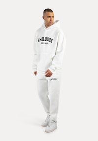 Biała bluza z kapturem i spodnie dresowe, z czarnym napisem "SMILODOX ATHL WEAR". Wykonana z miękkiego materiału, z przednią kieszenią i mankietami.