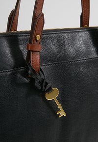 Zwarte leren tas met textuur, voorzien van bruine verstelbare hengsels en een gouden sleutelhanger eraan bevestigd. Ritssluiting. Eenvoudige hardware-accenten.