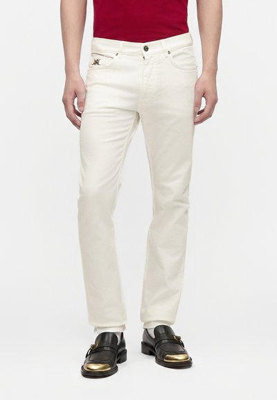 Homme portant un pantalon blanc slim, une chemise rouge et des chaussures en cuir noir avec des bouts dorés, debout devant un fond clair uni.