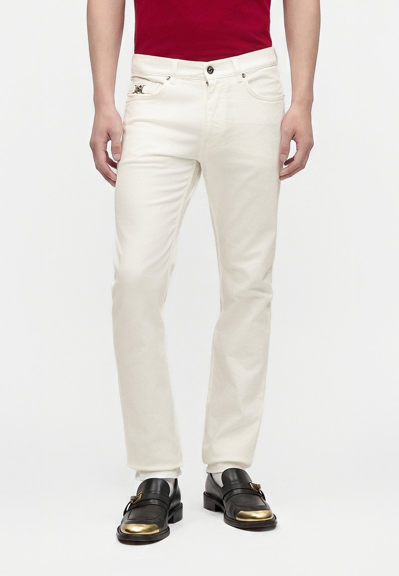 Homme portant un pantalon blanc slim, une chemise rouge et des chaussures en cuir noir avec des bouts dorés, debout devant un fond clair uni.