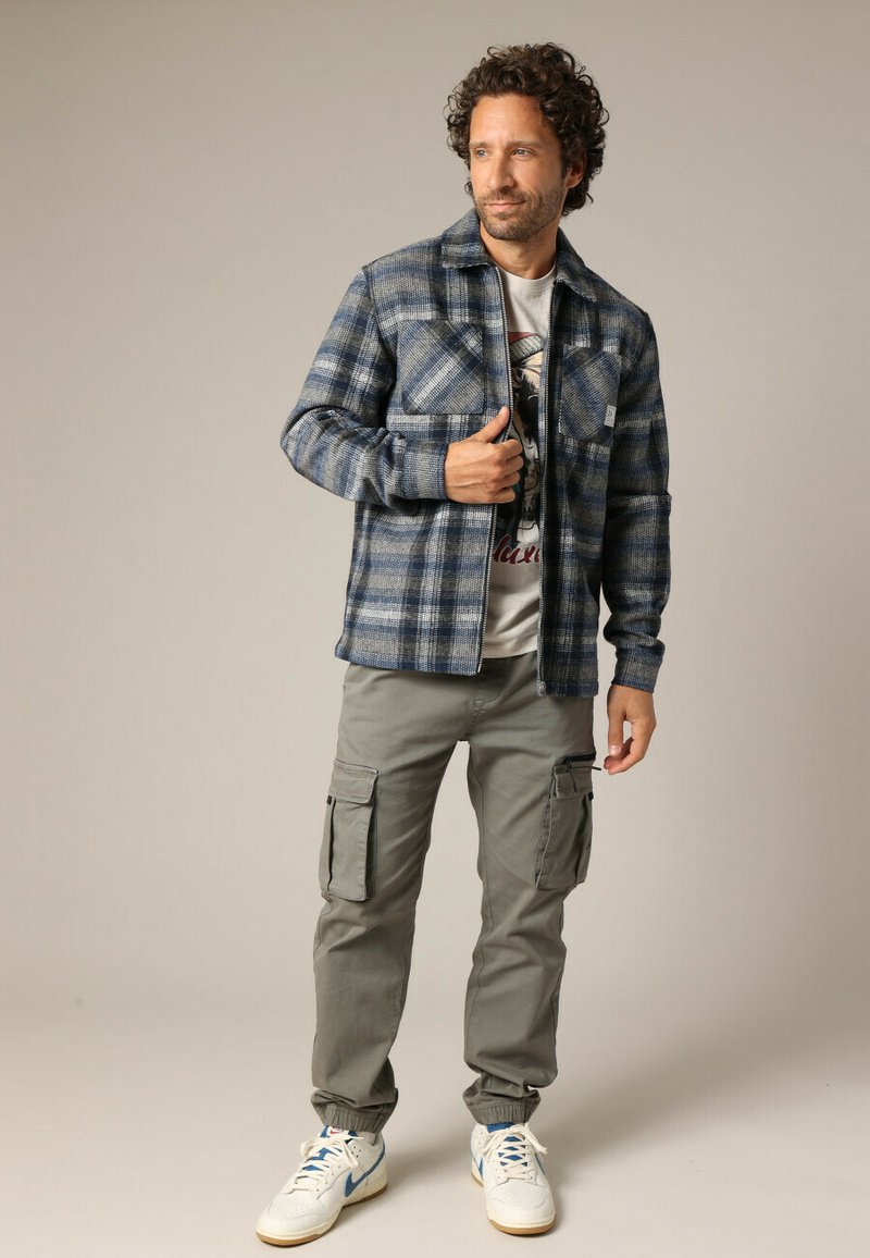 Blauw en grijs geruite flanellen shirt met twee borstzakken, beige graphic t-shirt eronder, grijze cargo broek en witte sneakers.