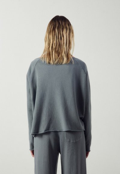 Personne aux cheveux blonds mi-longs, vue de dos, portant un haut gris ample à manches longues et un pantalon assorti sur un fond uni clair.