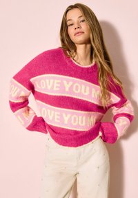 Rózsaszín kötött pulóver krémszínű "LOVE YOU" felirattal, hosszú ujjú és kerek nyakú. Világos színű nadrággal viselve, amely kis szívecskékkel díszített.