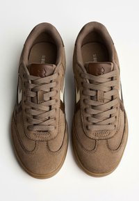 Brown suede sneakers with cream and dark brown accents. Rounded shape, lace-up front, and textured rubber sole. Clean design and stitching details.